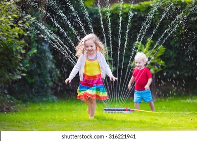 Child Playing With Garden Sprinkler. Preschooler Kid Running And Jumping. Summer Outdoor Water Fun In The Backyard. Children Play With Hose Watering Flowers. Kids Run And Splash On Hot Sunny Day.