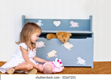 Child Playing With Her Doll While A Teddy Bear Pokes His Head Out Of The Toy Chest.