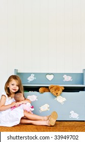 Child Playing With Her Doll While A Teddy Bear Pokes His Head Out Of The Toy Chest.