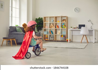 Child Playing At Home. Back View Of A Little Curly Girl In A Red Cloak And Helmet Rides A Tricycle At Home In The Room. Concept Of A Happy Childhood.