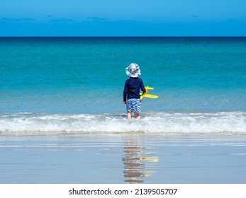 Child playing on the beach wearing wide brimmed hat, rashie or rash vest, sun protective clothing. Young Australian boy playing in the water with a toy aeroplane. Long sleeved top and board shorts - Shutterstock ID 2139505707