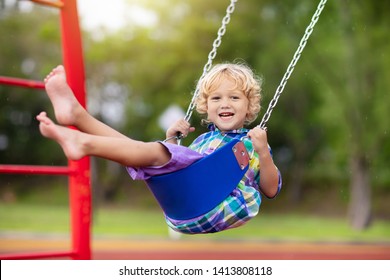 Child playing on outdoor playground in rain. Kids play on school or kindergarten yard. Active kid on colorful swing. Healthy summer activity for children in rainy weather. Little boy swinging.