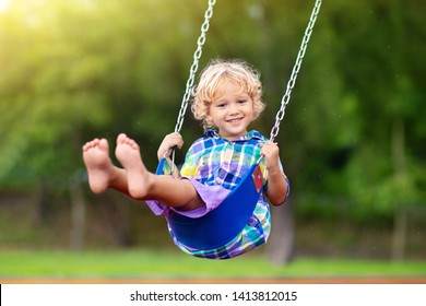 Child playing on outdoor playground in rain. Kids play on school or kindergarten yard. Active kid on colorful swing. Healthy summer activity for children in rainy weather. Little boy swinging.