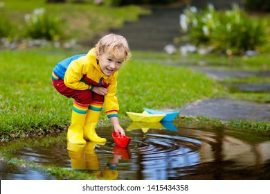 Child Playing With Paper Boat In Puddle. Kids Play Outdoor By Autumn Rain. Fall Rainy Weather Outdoors Activity For Young Children. Kid Jumping In Muddy Puddles. Waterproof Jacket And Boots For Baby.