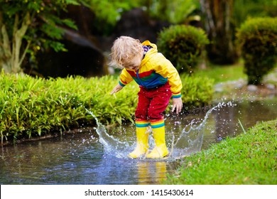 Child Playing In Puddle. Kids Play And Jump Outdoor By Autumn Rain. Fall Rainy Weather Outdoors Activity For Young Children. Kid Jumping In Muddy Puddles. Waterproof Jacket And Boots For Little Boy.