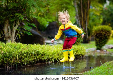 Child Playing In Puddle. Kids Play And Jump Outdoor By Autumn Rain. Fall Rainy Weather Outdoors Activity For Young Children. Kid Jumping In Muddy Puddles. Waterproof Jacket And Boots For Little Boy.