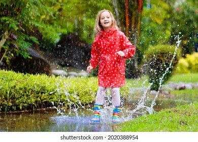 Child Playing In Puddle. Kids Play And Jump Outdoor By Autumn Rain. Fall Rainy Weather Outdoors Activity For Young Children. Kid Jumping In Muddy Puddles. Waterproof Jacket And Boots For Little Girl.