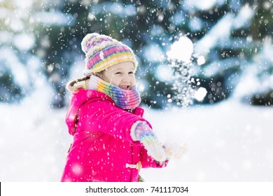 Child Playing With Snow In Winter. Little Girl In Colorful Jacket And Knitted Hat Catching Snowflakes In Winter Park On Christmas. Kids Play And Jump In Snowy Forest. Snow Ball Fight For Children.