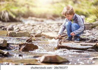 Child Playing In Summer Sunshine With A Paper Boat In Stream