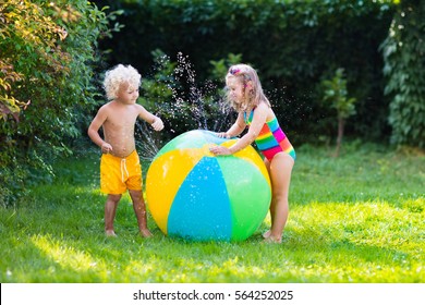 Child Playing With Toy Ball Garden Sprinkler. Preschooler Kid Run And Jump. Summer Outdoor Water Fun In The Backyard. Children Play With Hose Watering Flowers. Kids Splash On Sunny Day