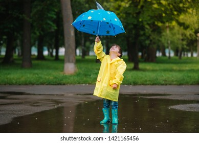 Child Playing With Toy Boat In Puddle. Kid Play Outdoor By Rain. Fall Rainy Weather Outdoors Activity For Young Children. Kid Jumping In Muddy Puddles. Waterproof Jacket And Boots For Baby. 