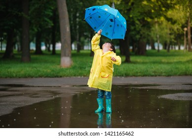 Child playing with toy boat in puddle. Kid play outdoor by rain. Fall rainy weather outdoors activity for young children. Kid jumping in muddy puddles. Waterproof jacket and boots for baby. 