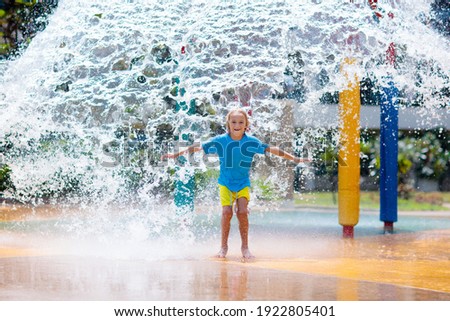 Child playing under tip bucket in water park. Kids play with splash dump bucket. Family fun in amusement center on hot summer day. Sun protection for children. Water slides for kid.