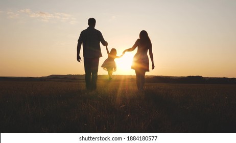 Child Plays With Dad And Mom On Field In Sunset Light. Family And Childhood. Little Daughter Jumping Holding Hands Of Dad And Mom In Park On Background Of Sun. Walking With A Small Kid In Nature.