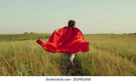 Child plays and dreams. Happy superhero girl, runs on green field in red cloak, cloak flutters in wind. Teenager dreams of becoming superhero. Young girl in red cloak, dream expression. - Shutterstock ID 2019852458