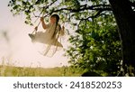 Child plays on wooden swing, dreams of flying. Baby swing, kid girl smile in flight. Happy little girl swings on swing in park under tree at sunset. Concept of family holiday, dreams, entertainment.