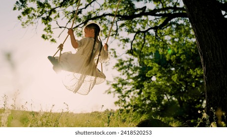 Child plays on wooden swing, dreams of flying. Baby swing, kid girl smile in flight. Happy little girl swings on swing in park under tree at sunset. Concept of family holiday, dreams, entertainment. - Powered by Shutterstock