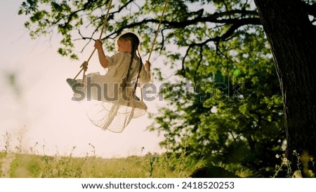 Child plays on wooden swing, dreams of flying. Baby swing, kid girl smile in flight. Happy little girl swings on swing in park under tree at sunset. Concept of family holiday, dreams, entertainment.