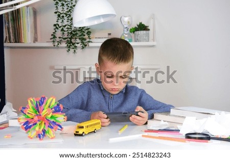 Child plays with smartphone instead of doing homework. Boy sits at desk and uses a mobile phone instead of reading a book. Education, school, learning disability, reading difficulties.