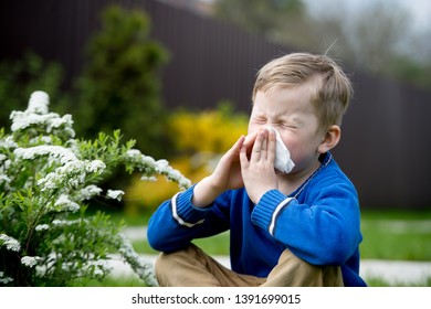 Child with pollen allergy. Boy sneezing and blowing nose because of seasonal allergy while sitting in a grass. Spring allergy concept. Flowering bushes and trees in background. Child allergy - Powered by Shutterstock