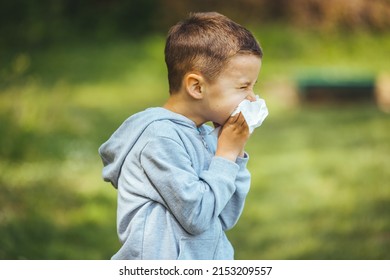 Child with pollen allergy. Boy sneezing and blowing nose because of seasonal allergy while sitting in a grass. Spring allergy concept. Flowering bushes and trees in background. Child allergy - Powered by Shutterstock
