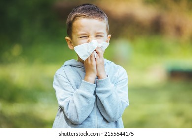 Child with pollen allergy. Boy sneezing and blowing nose because of seasonal allergy while sitting in a grass. Spring allergy concept. Flowering bushes and trees in background. Child allergy