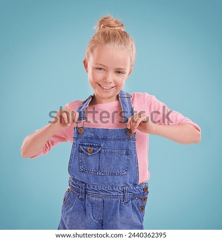 Child, portrait and smile in studio with confidence in dungarees on blue background, fashion or mockup space. Girl, school student and positive childhood in Australia with good mood, growth or kid