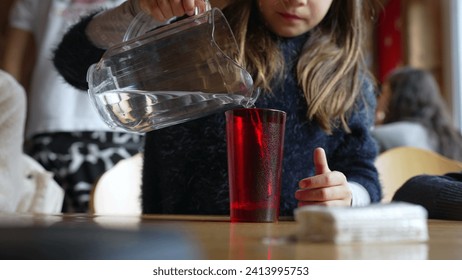 Child pouring water into red plastic cup with jar standing at restaurant, small girl serving refreshing tap water at diner - Powered by Shutterstock