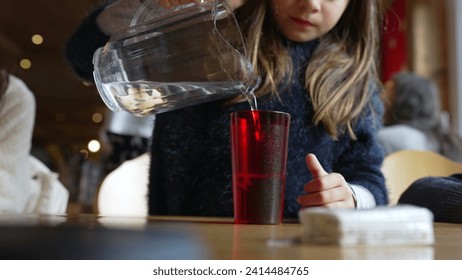 Child pouring water into red plastic cup with jar standing at restaurant, small girl serving refreshing tap water at diner - Powered by Shutterstock