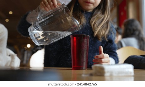 Child pouring water into red plastic cup with jar standing at restaurant, small girl serving refreshing tap water at diner - Powered by Shutterstock