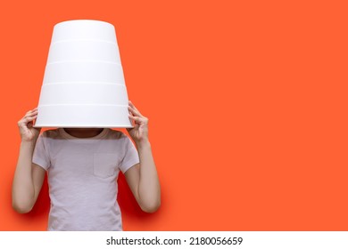 The child put a bucket on his head, sitting on an orange background. Funny child hid in a white bucket. The child hides from the parents. Free space for text