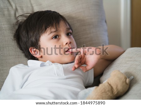 Child putting finger in his mouth.Schoolboy biting his finger nails while watching TV, Emotional kid portrait, Young boy siting on sofa looking out with thinking face or nervous, Children Health care 