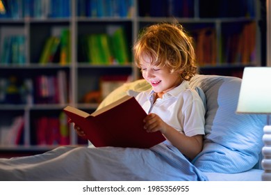 Child Reading Book In Bed. Kids Read At Night. Little Boy With Fairy Tale Books In Bedroom . Education For Young Children. Bedtime Story In The Evening. Cute Kid Under Blanket In Dark Room With Lamp.