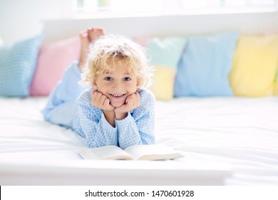 Child Reading Book In Bed In White Sunny Bedroom With Window. Children Read Books. Kids Room. Little Blond Curly Boy In Pajamas At Home. Bedding And Sleepwear For Nursery. Kid With Toy.