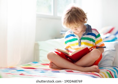 Child Reading Book In Bed In White Sunny Bedroom With Window. Children Read Books. Kids Room. Little Blond Curly Boy In Pajamas At Home. Bedding And Sleepwear For Nursery. Kid With Toy.