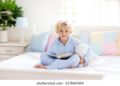 Child reading book in bed in white sunny bedroom with window. Children read books. Kids room. Little blond curly boy in pajamas at home. Bedding and sleepwear for nursery. Kid with toy.