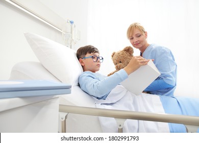 Child Reading A Book And Lying In Bed In Hospital Room With A Nurse Holding A Teddy Bear Sitting Beside Him