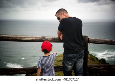 A child in red cap standing behind fence, talking to a man in black t-shirt. Dad talking, teaching his son relationship facing the sea on cloudy rainy day.