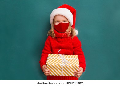 Child In A Red Sweater And A Santa Hat Wearing Medical Face Mask With Christmas Gift Box. Christmas Shopping.  Copy Space. 