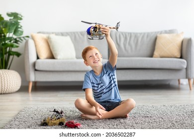 Child Redhead Boy Playing With Toys Alone At Home. Kids Play Concept. Cheerful Boy Sitting On Carpet In Cozy Living Room, Playing With Helicopter And Cars, Enjoying His Brand New Toys, Copy Space