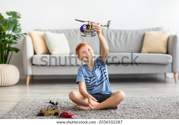 Child redhead boy playing with toys alone at home.\
Kids play concept. Cheerful boy sitting on carpet in cozy living\
room, playing with helicopter and cars, enjoying his brand new\
toys, copy space