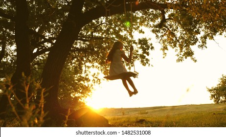 Child Rides A Rope Swing On An Oak Branch In Forest. Girl Laughs, Rejoices. Young Girl Swinging On Swing Under A Tree In Sun, Playing With Children. Close-up. Family Fun In Park, In Nature.