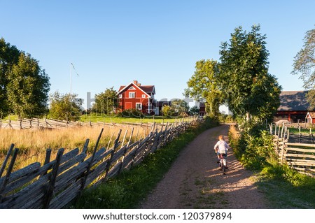 Child riding a bicycle along a small farm lane in rural Sweden pedaling towards a traditional red painted timber house