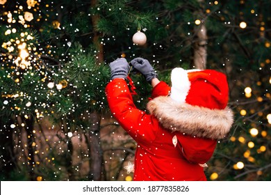 Child In Santa Claus Hat And Red Jacket Is Hanging Christmas Decorations On A Fir-tree In Theforest.