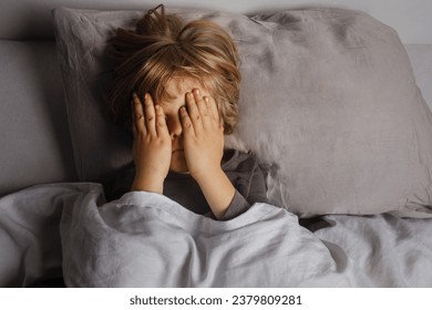 The child was scared before going to bed. Night terrors in a child. The kid covers his face with his hands in a fear. Children's experiences. Boy in the bed. View from above. Sad psychological state. 