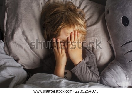 The child was scared before going to bed. Night terrors in a child. The kid covers his face with his hands in a fear. Children's experiences. Boy in the bed. Sad psychological state. 
Terrible dream.