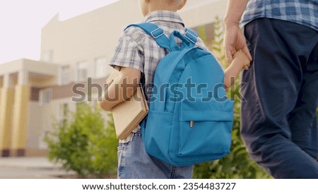 Child Schoolboy walking down street with backpack, holding his dad by hand. Happy family, father, child go to school together. Dad is taking his little son with textbook to school. Preschool education