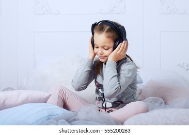 Child Schoolgirl Sitting On A Bed And Listening To An Audiobook. The Concept Of Childhood, Education And Music