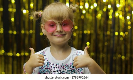 Child show thumbs up, smiling, looking at camera, demonstrating approval, satisfied with excellent result. Little fun blonde kid teen girl 4-5 years old in pink sunglasses posing on shiny background