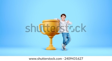 Child showing a thumb up, full length standing near big gold champion cup on light blue background. Concept of first place, winner and success in sports